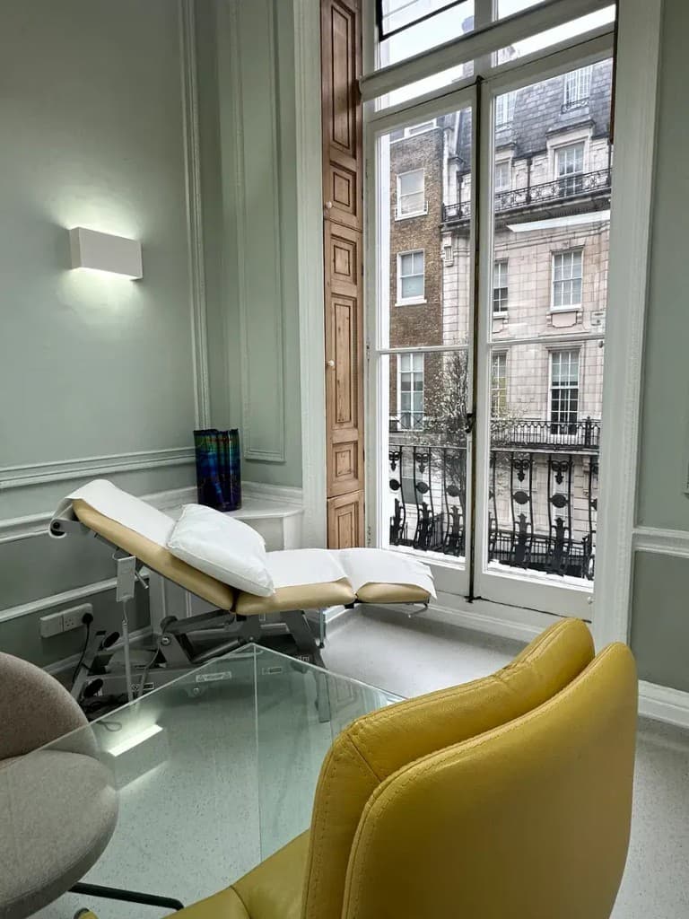 Treatment room with a consultation chair and bed beside a tall window